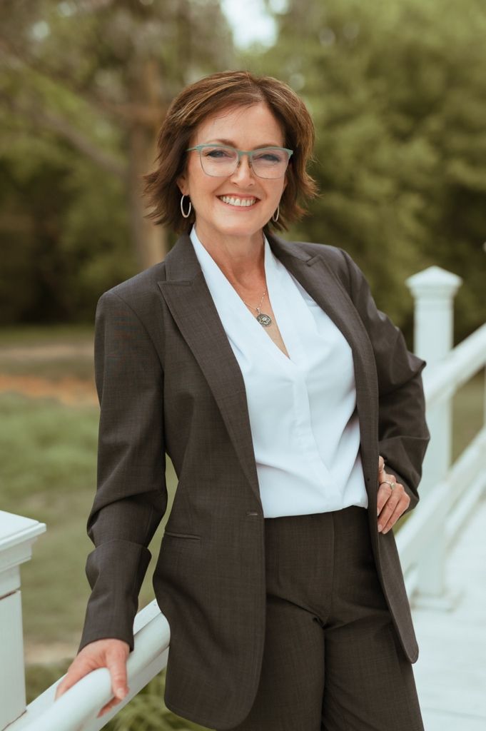 Professional wearing a brown business suit and glasses poses confidently on white porch railing outdoors.