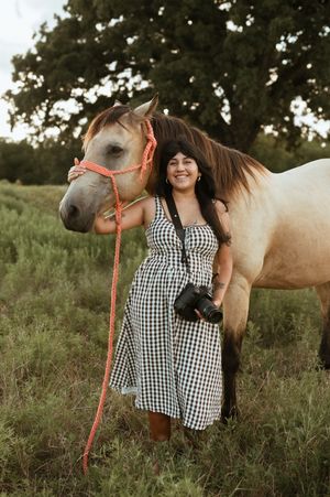 A beautiful cream-colored horse with orange halter stands in a grassy field with a woman in a gingham dress at sunset.