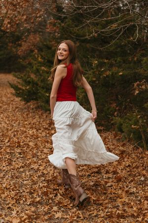 Person in red top and white dress twirls playfully among fallen autumn leaves with brown boots in a forest setting.
