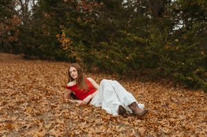 A person in a red top and white skirt lies on autumn leaves in a forest setting during fall.
