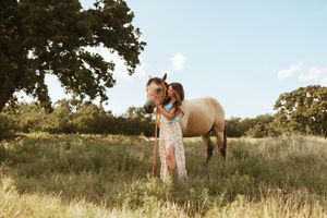 A white dress-wearing person stands in a sunlit meadow with a brown horse on a warm summer day.