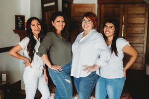 Four women stand together casually posing and smiling in jeans and casual tops against a wooden door.