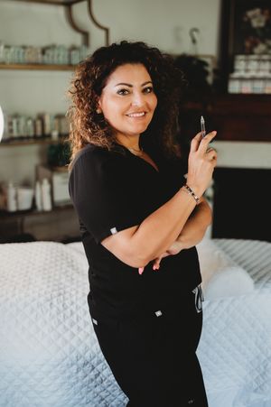 Healthcare professional in black scrubs stands confidently with curly hair in a medical spa or treatment room setting.