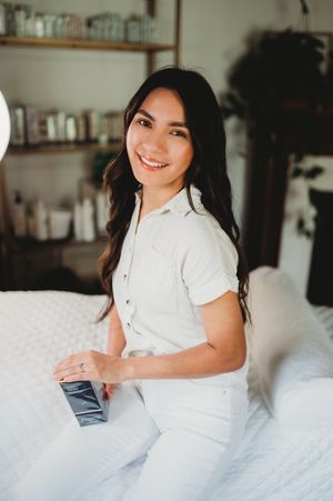 Smiling young woman in white shirt sits on a bed with wooden shelves full of products in the background.