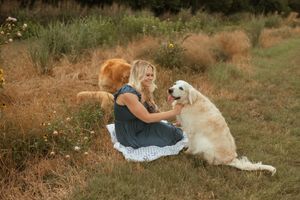 A golden retriever and someone sit together on a blanket in a rustic outdoor setting with tall grass.