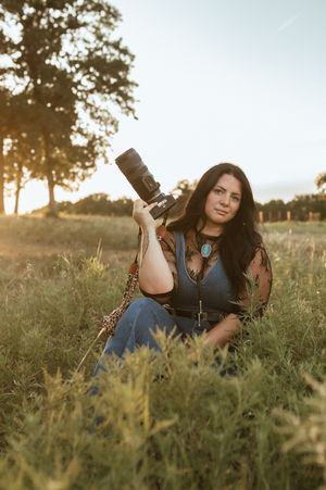 Professional photographer in blue dress lying in grassy field at sunset holding camera lens and smiling.