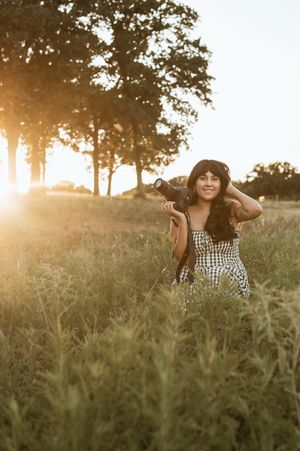 A person in a checkered dress sits in a grassy field at sunset while sunlight streams through trees in the background.