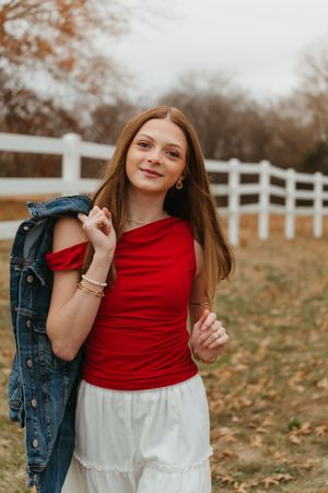 Young person in a red top and white skirt poses playfully outdoors near a white fence on an autumn day.