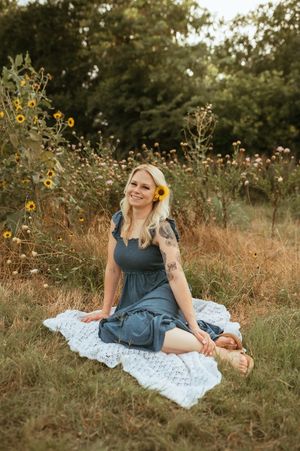 Someone in a navy blue dress sits on a white blanket surrounded by yellow wildflowers in a summer field.