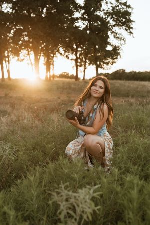 A portrait in a sundress crouching in a grassy field at sunset while trees silhouette against the golden sky.