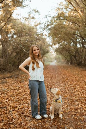 A person and their puppy sit together on a leaf-covered autumn path surrounded by trees.