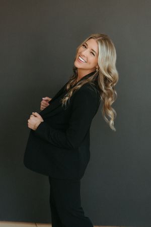 Smiling blonde woman in a black blazer and pants poses sideways against a dark gray background.