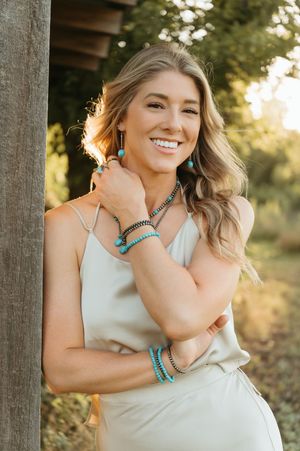 Smiling blonde woman wearing turquoise jewelry, bracelets, and earrings with a white dress, leaning against a wooden post outdoors.