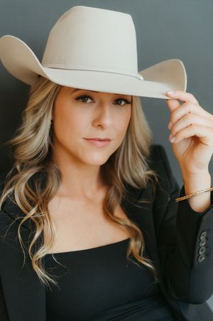 Blonde woman in black blazer poses confidently, tipping a wide-brim beige cowboy hat, wearing a gold bracelet.