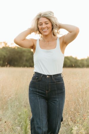 Smiling blonde woman in white tank top and dark jeans poses in golden field at sunset.