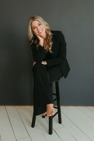 Smiling blonde woman in black blazer and pants sitting on a stool against a dark gray wall.