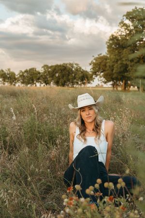 Woman in white cowboy hat sitting in a wildflower meadow with trees and cloudy sky backdrop.