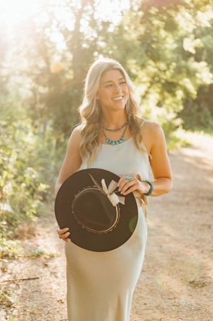 Smiling blonde woman in a white dress holding a black hat, wearing turquoise jewelry, standing on a sunny outdoor path.
