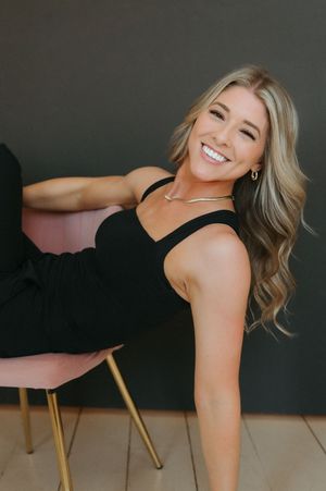 Smiling blonde woman in black outfit relaxing on a pink and gold chair against a dark gray background.
