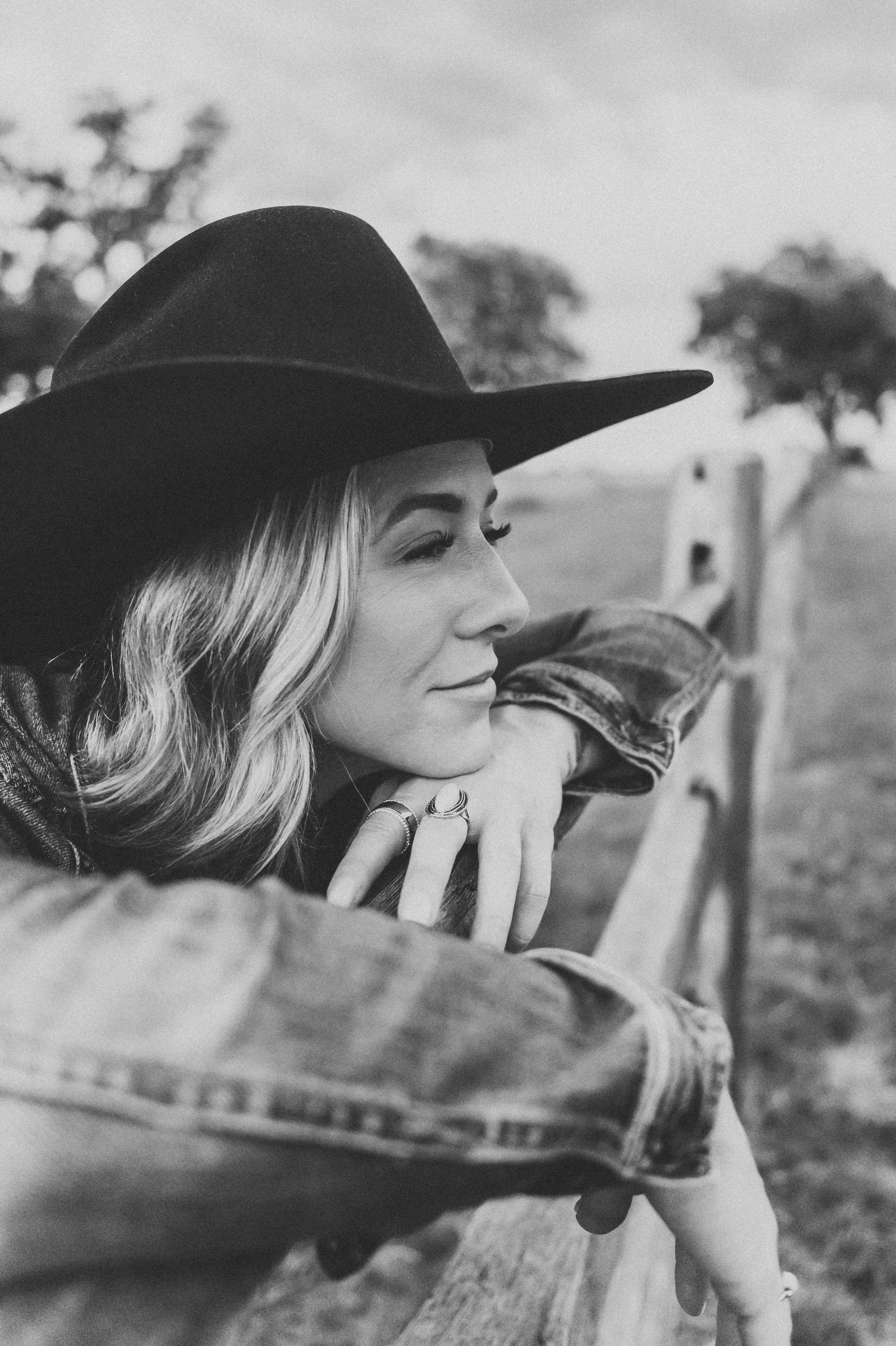 Black and white portrait of blonde woman in cowboy hat leaning on rustic wooden fence outdoors.