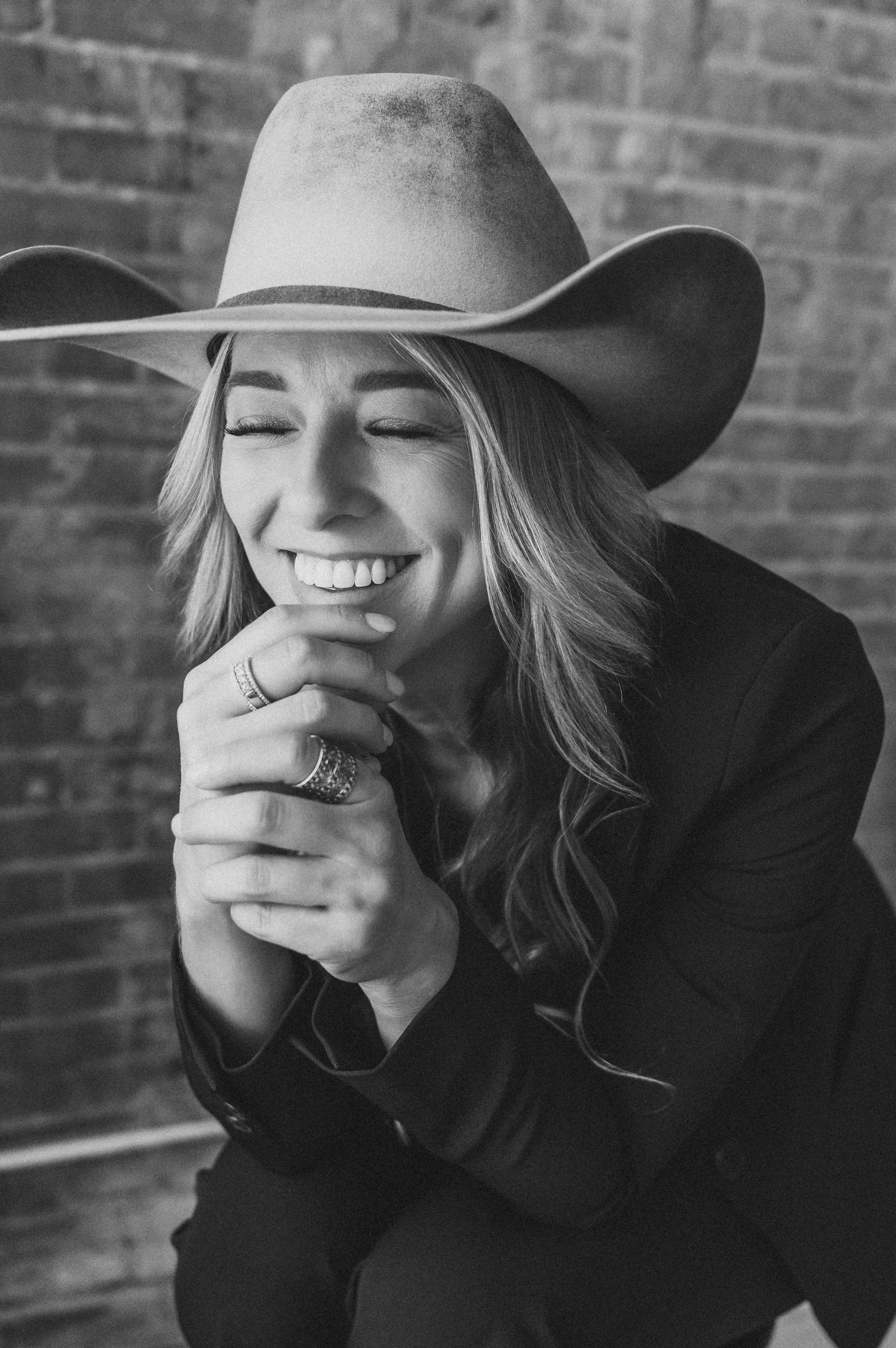 Black and white photo of a smiling blonde woman in a wide-brim hat, hands clasped, against a brick wall.