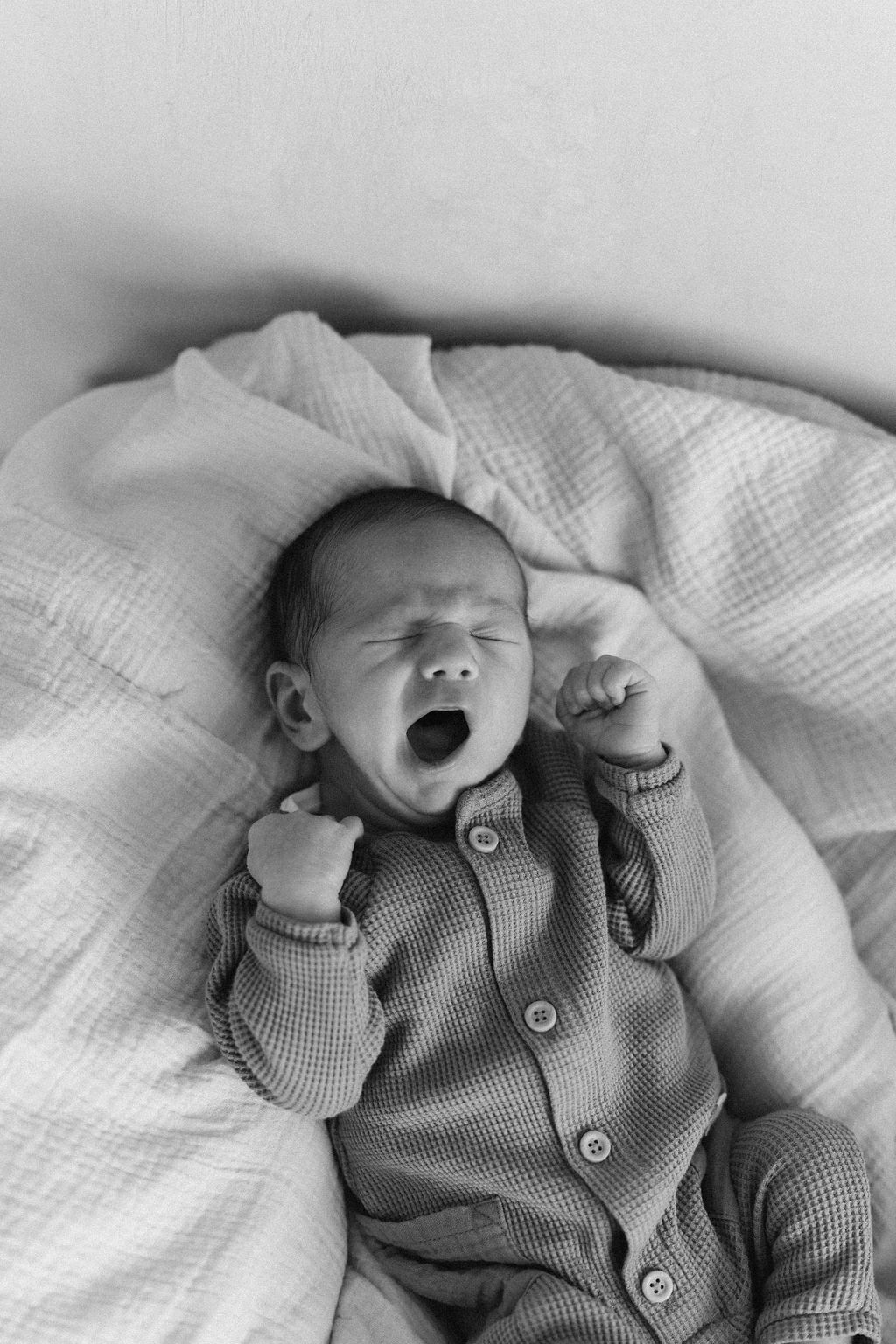 Sweet newborn in gray buttoned sleeper yawning while lying on white blanket in black and white photograph.