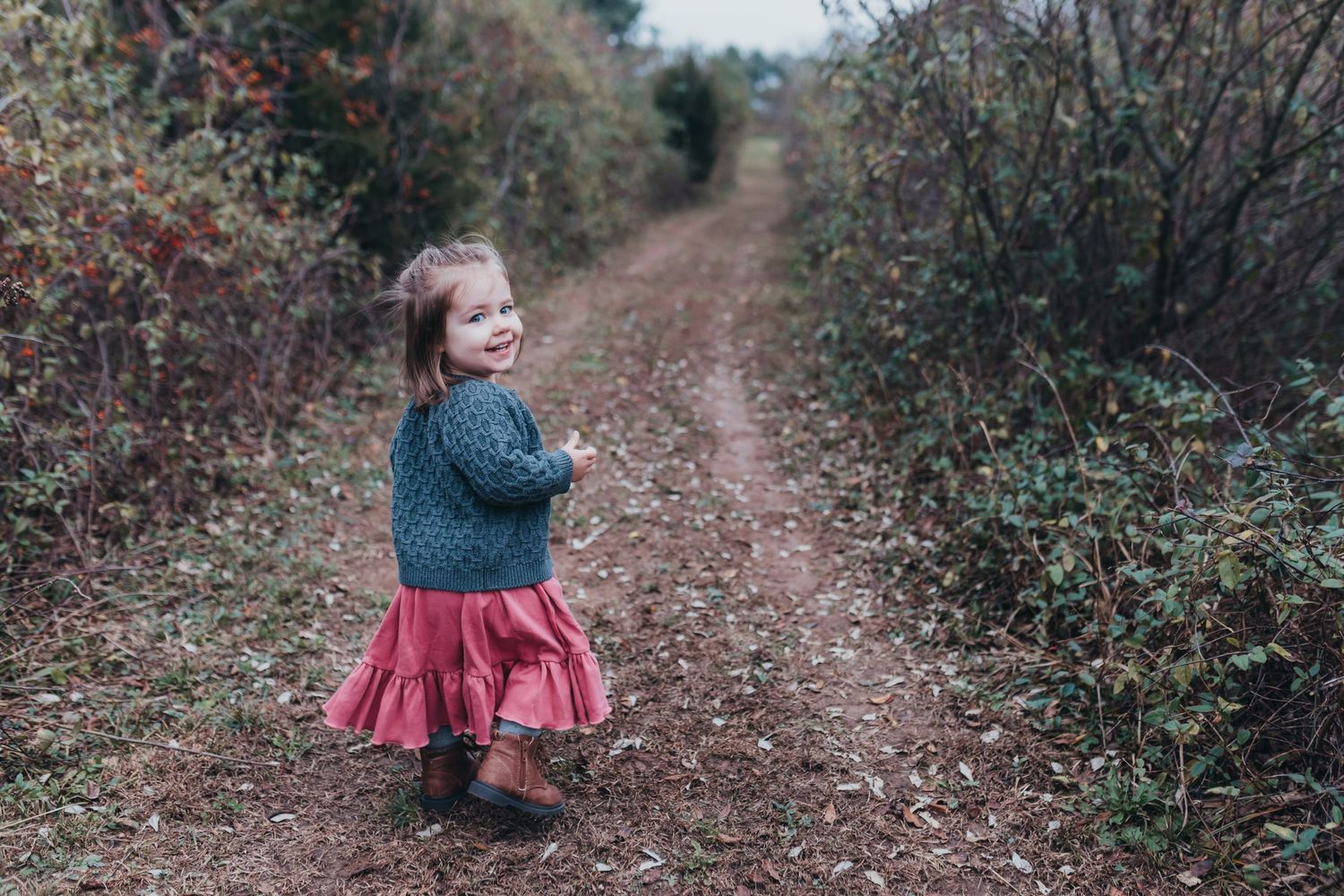 Small child in a twirly red skirt exploring a dirt path through autumn bushes.