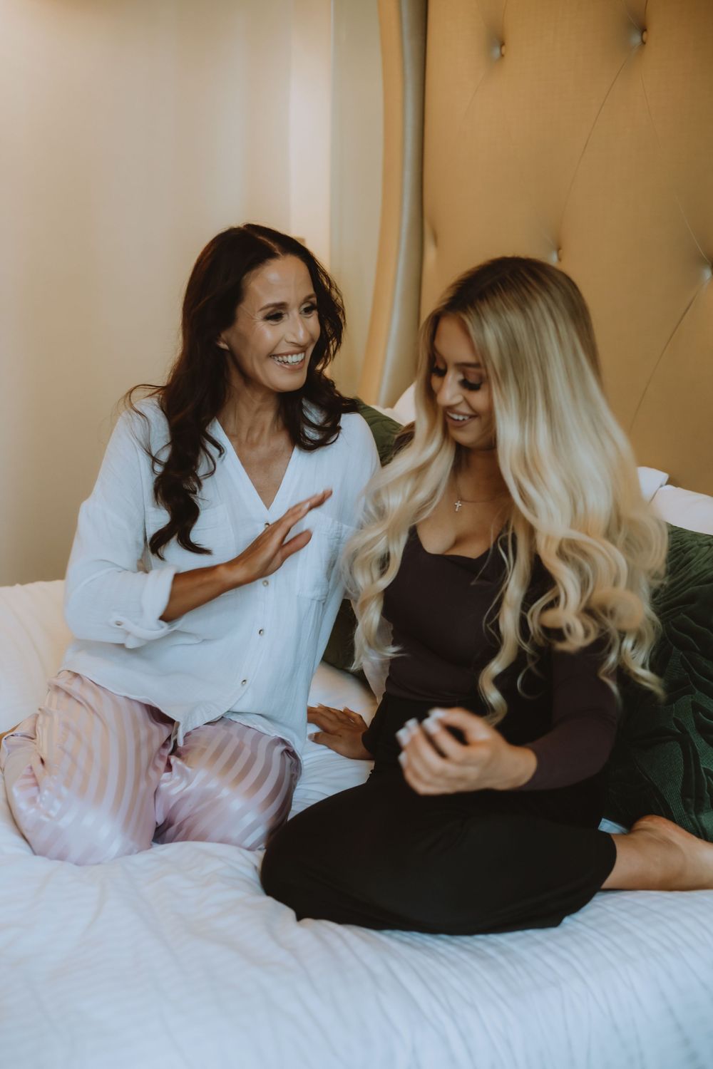 Two friends sharing a joyful moment while sitting on a white bed, one wearing brown and one in white.