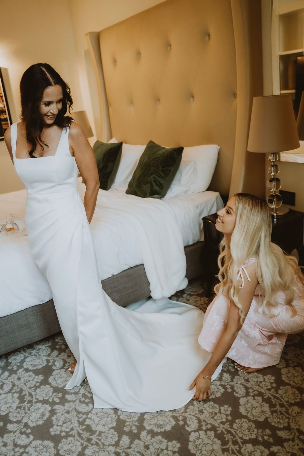 A bride sits on a gray couch in a flowing white wedding dress against a floral patterned carpet.