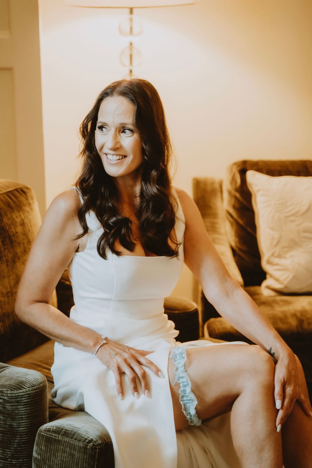 A woman in a white dress smiles warmly while sitting comfortably on a couch in a soft-lit room.