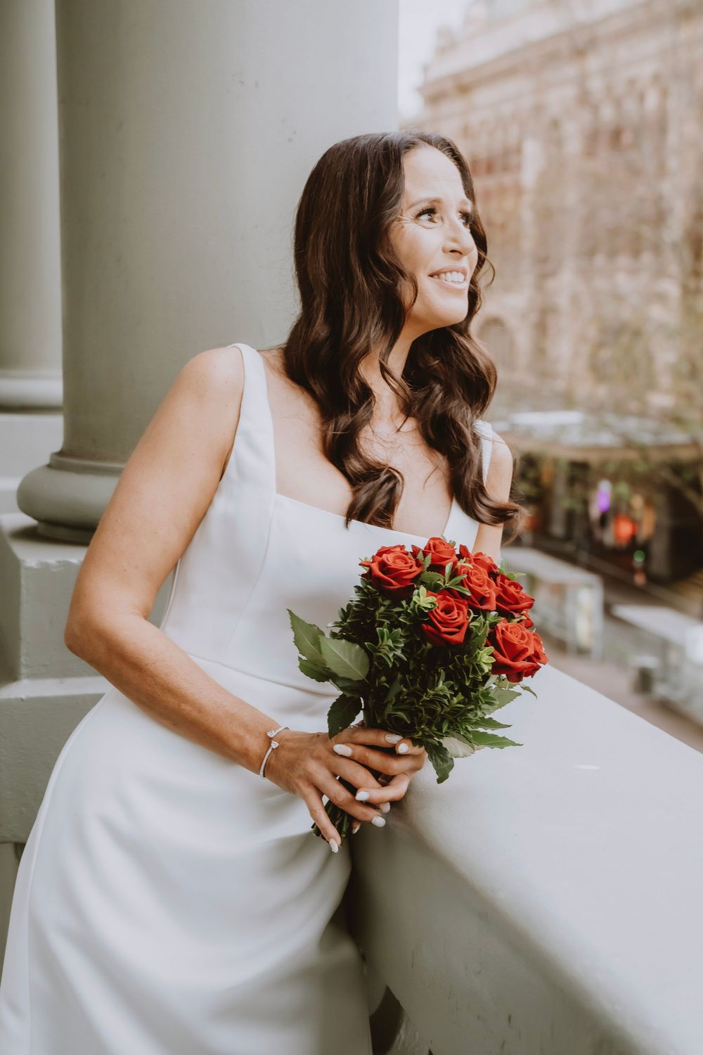 A bride in a white dress holds a bouquet of red roses while smiling and leaning against a balcony railing.