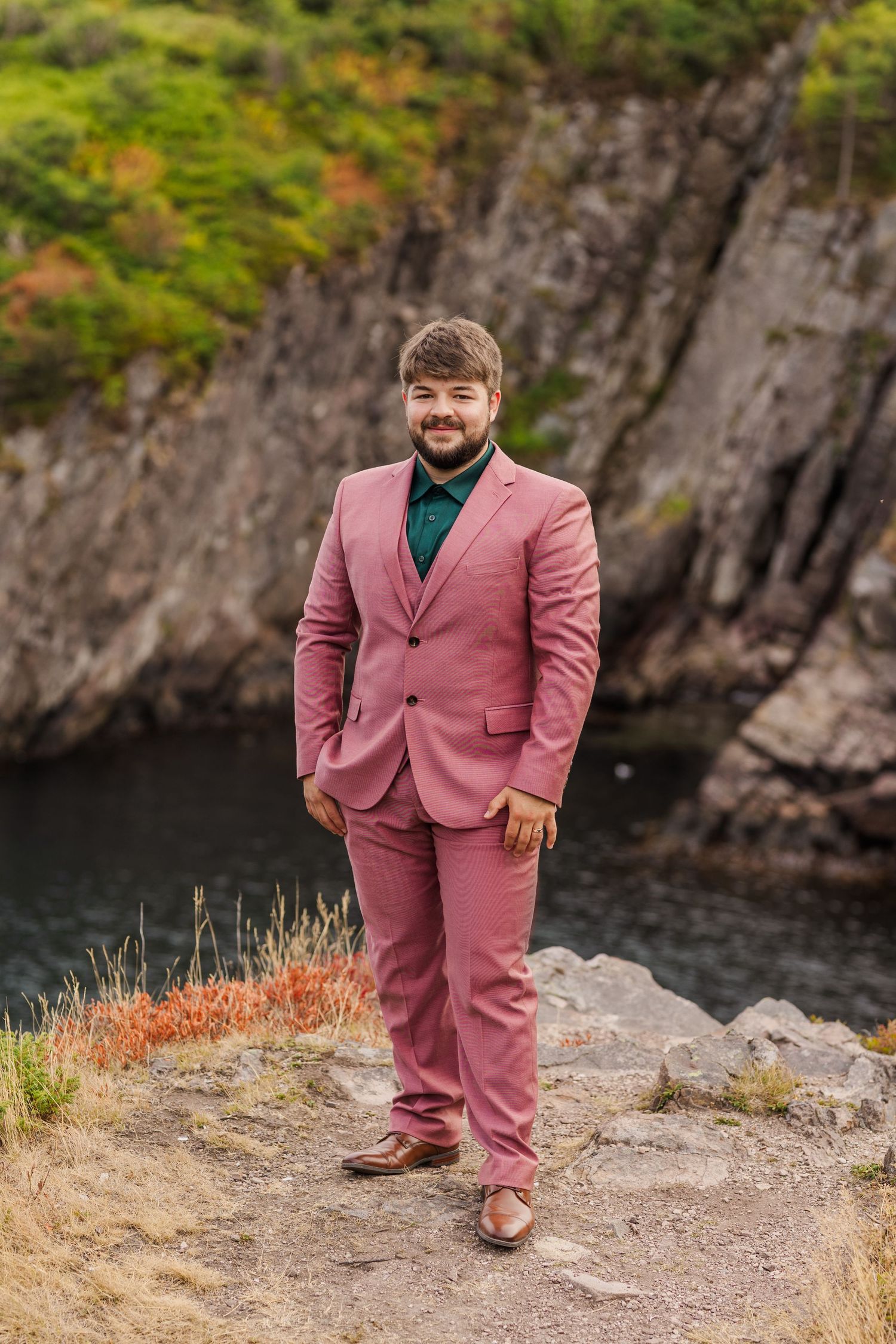 A person wearing a pink suit poses outdoors near rocky cliffs and water in autumn scenery.