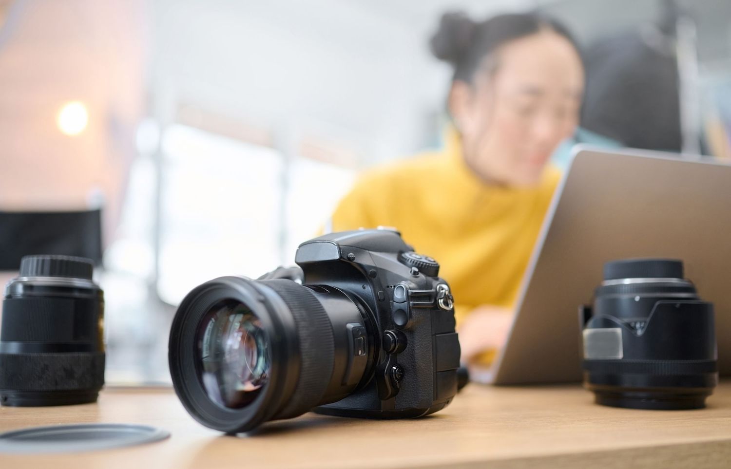 Professional DSLR camera with lens on desk next to laptop with additional camera lens in foreground.