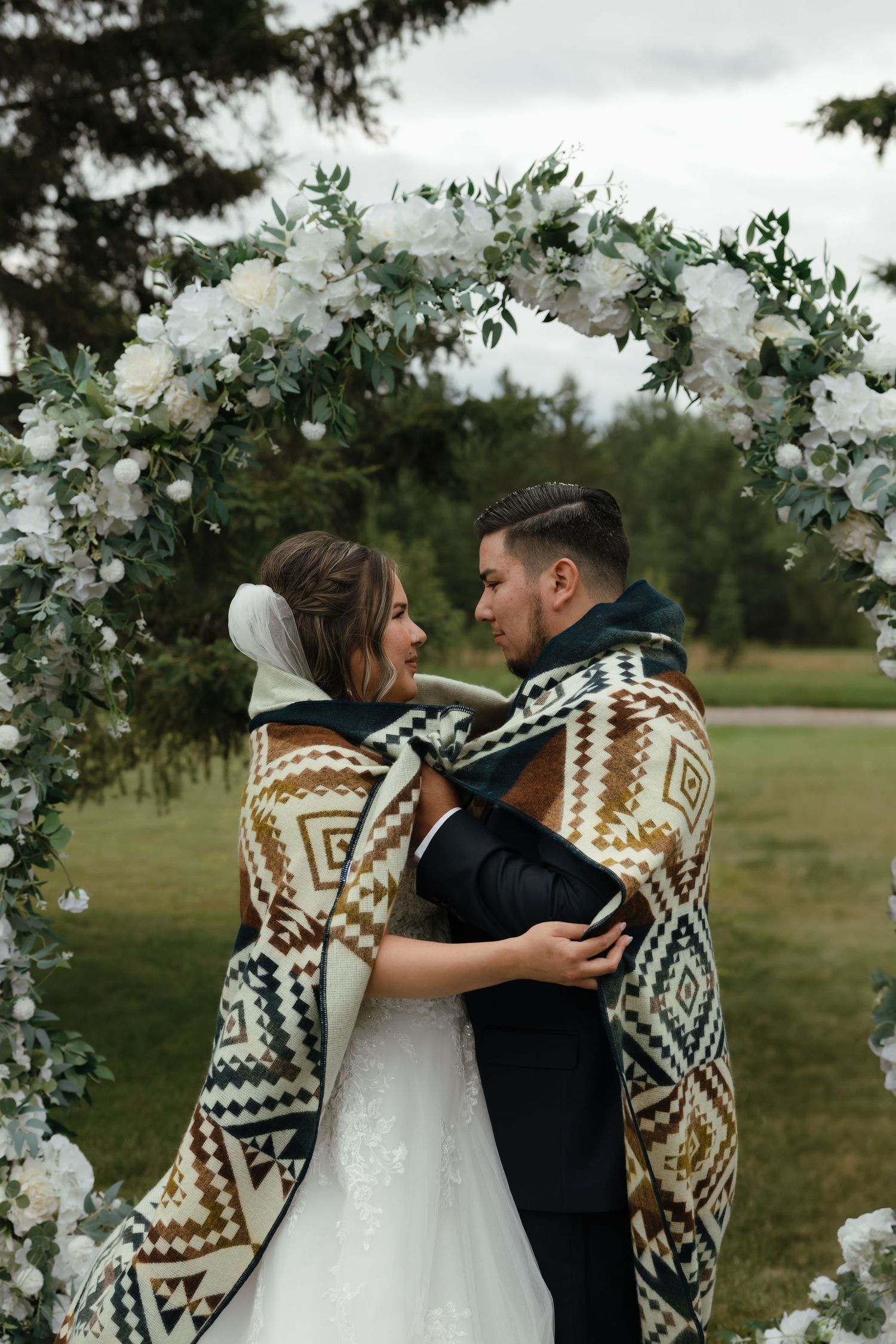 A couple wrapped in geometric patterned blankets embrace under a floral arch in an outdoor wedding setting.