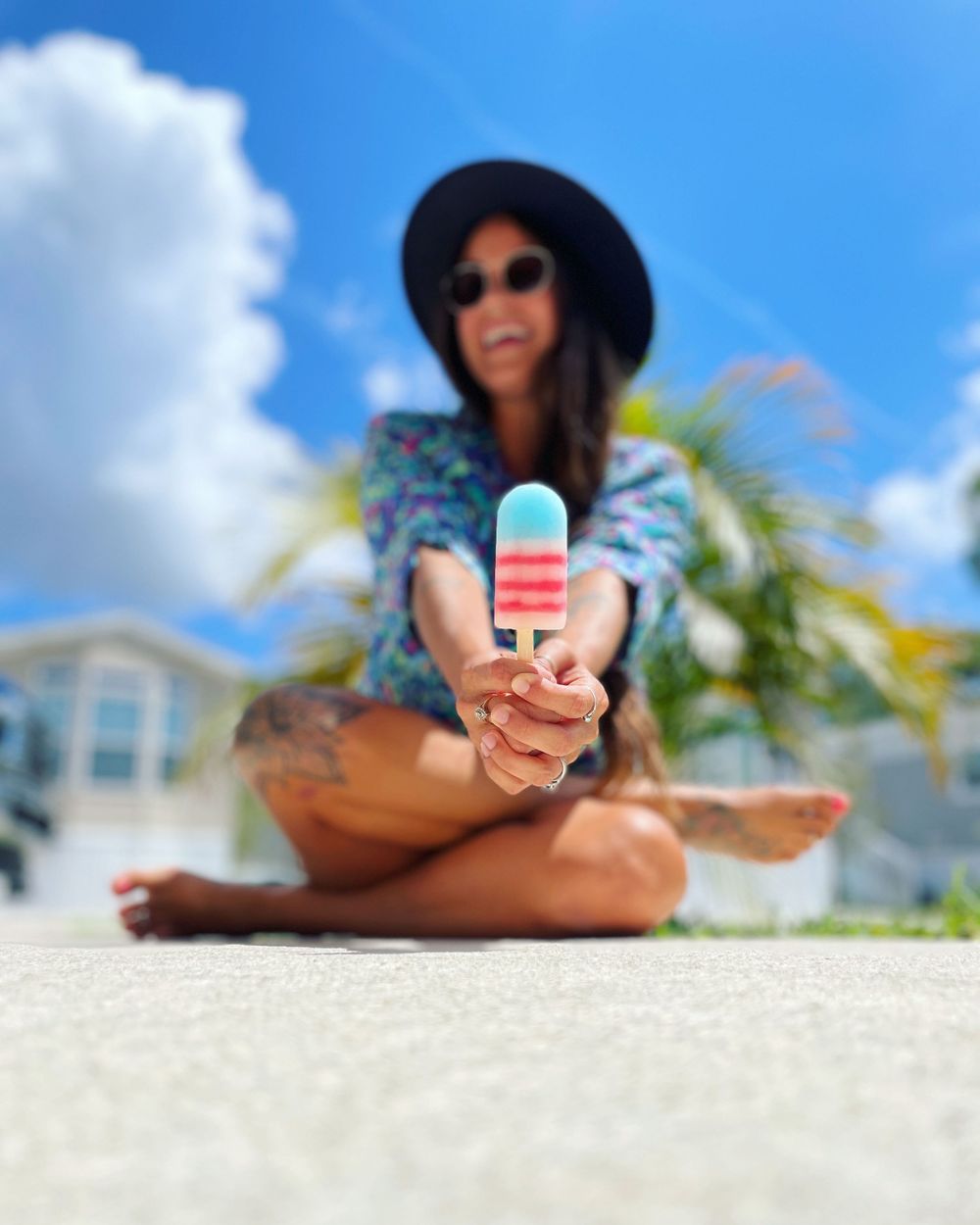 A tattooed woman in a black hat sits cross-legged outdoors holding a red white and blue popsicle toward the camera.