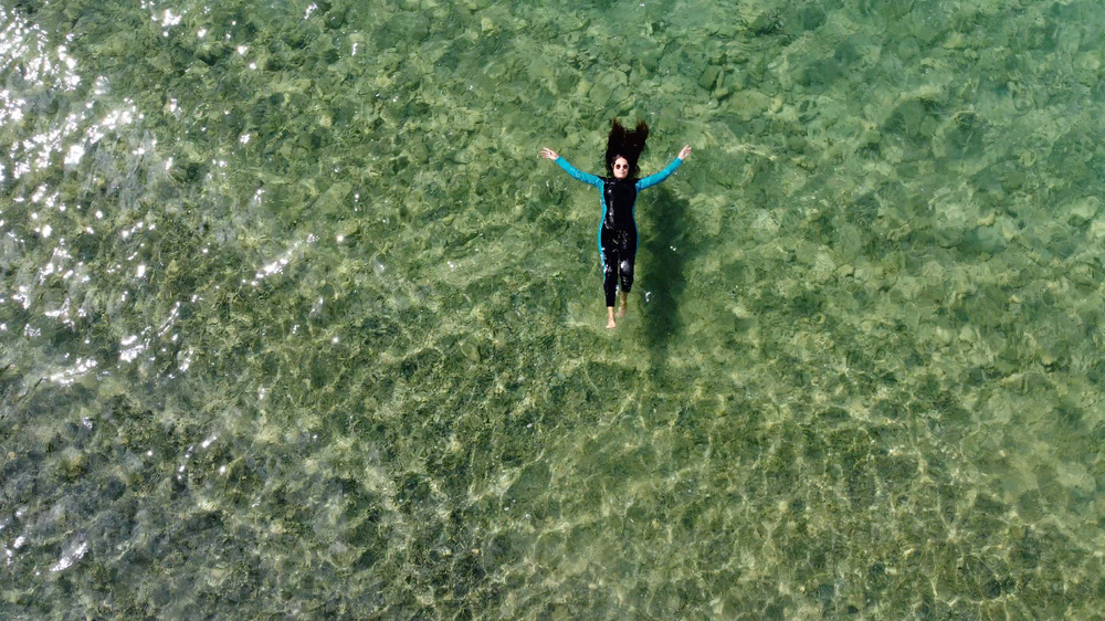 Aerial view of a person in a wetsuit floating spread-eagle in shallow clear green ocean water.