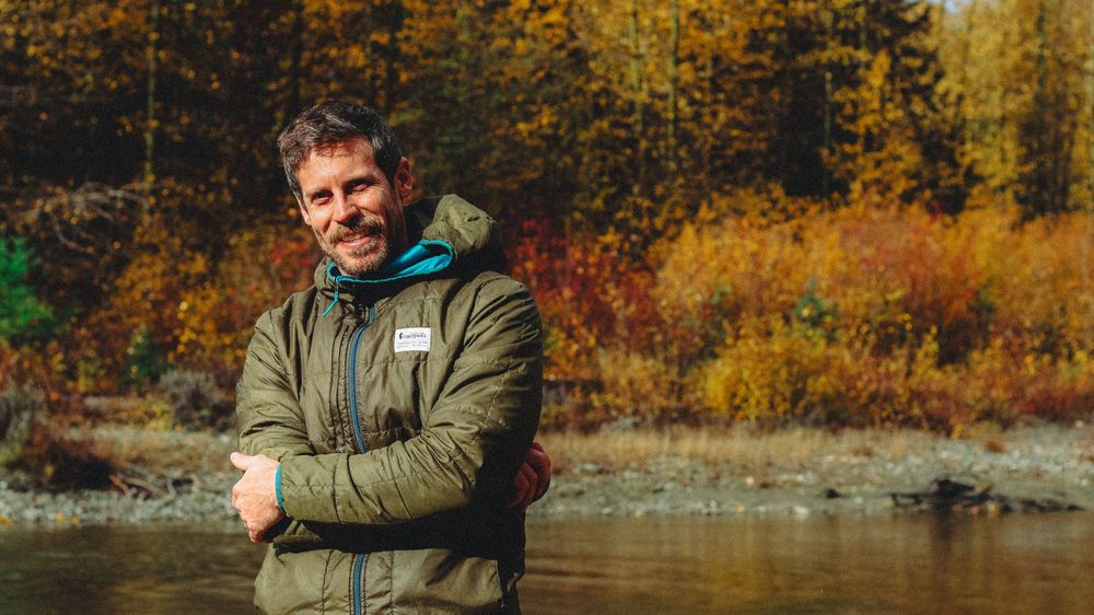 Smiling man in green jacket standing by river with vibrant autumn foliage background.