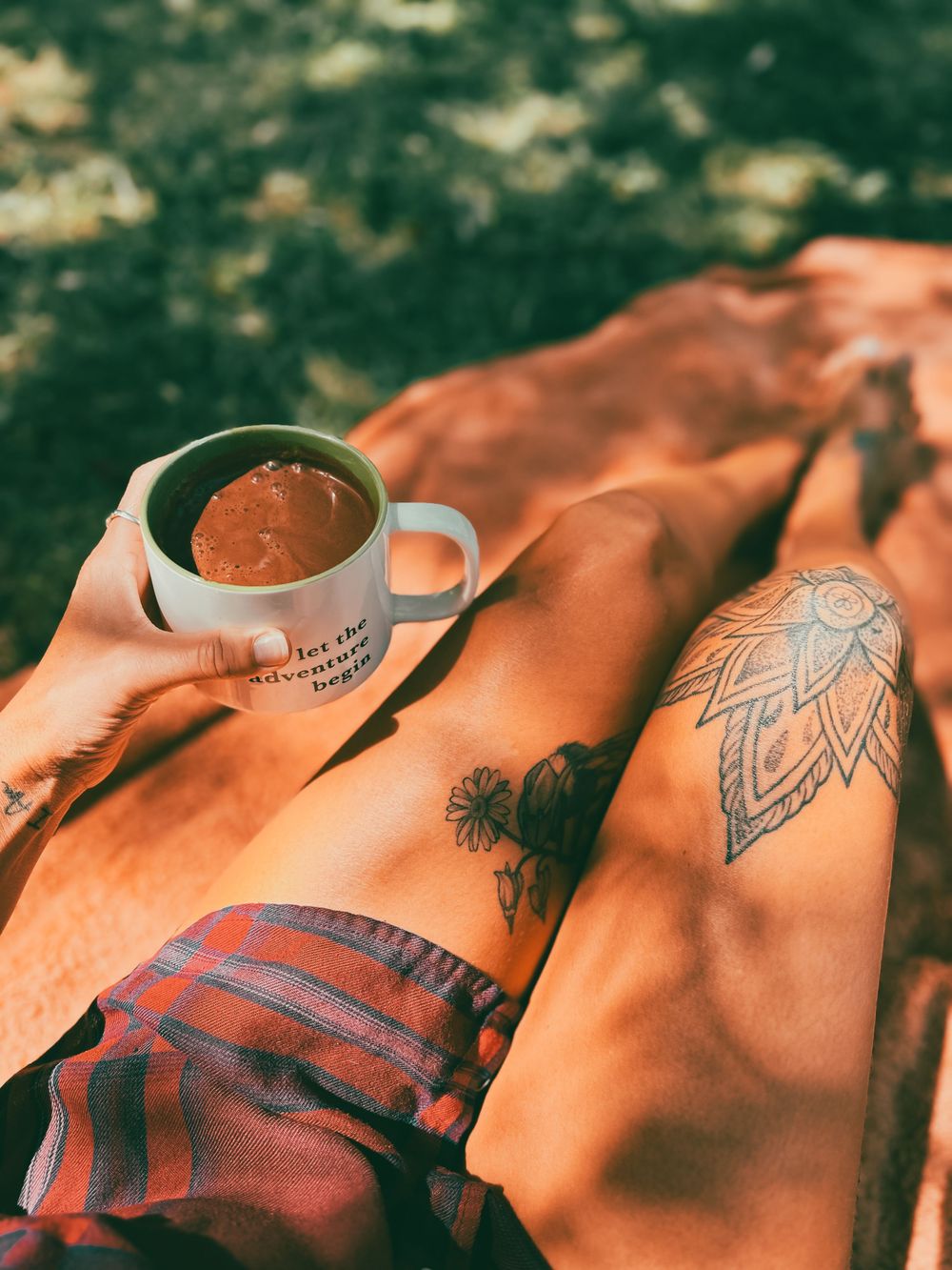 Person relaxing outdoors holding adventure mug with mandala tattoos visible on legs on orange blanket.