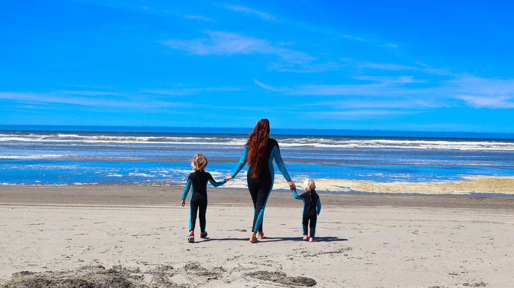 A mother holds hands with two young children in wetsuits walking toward the ocean on a sunny beach.