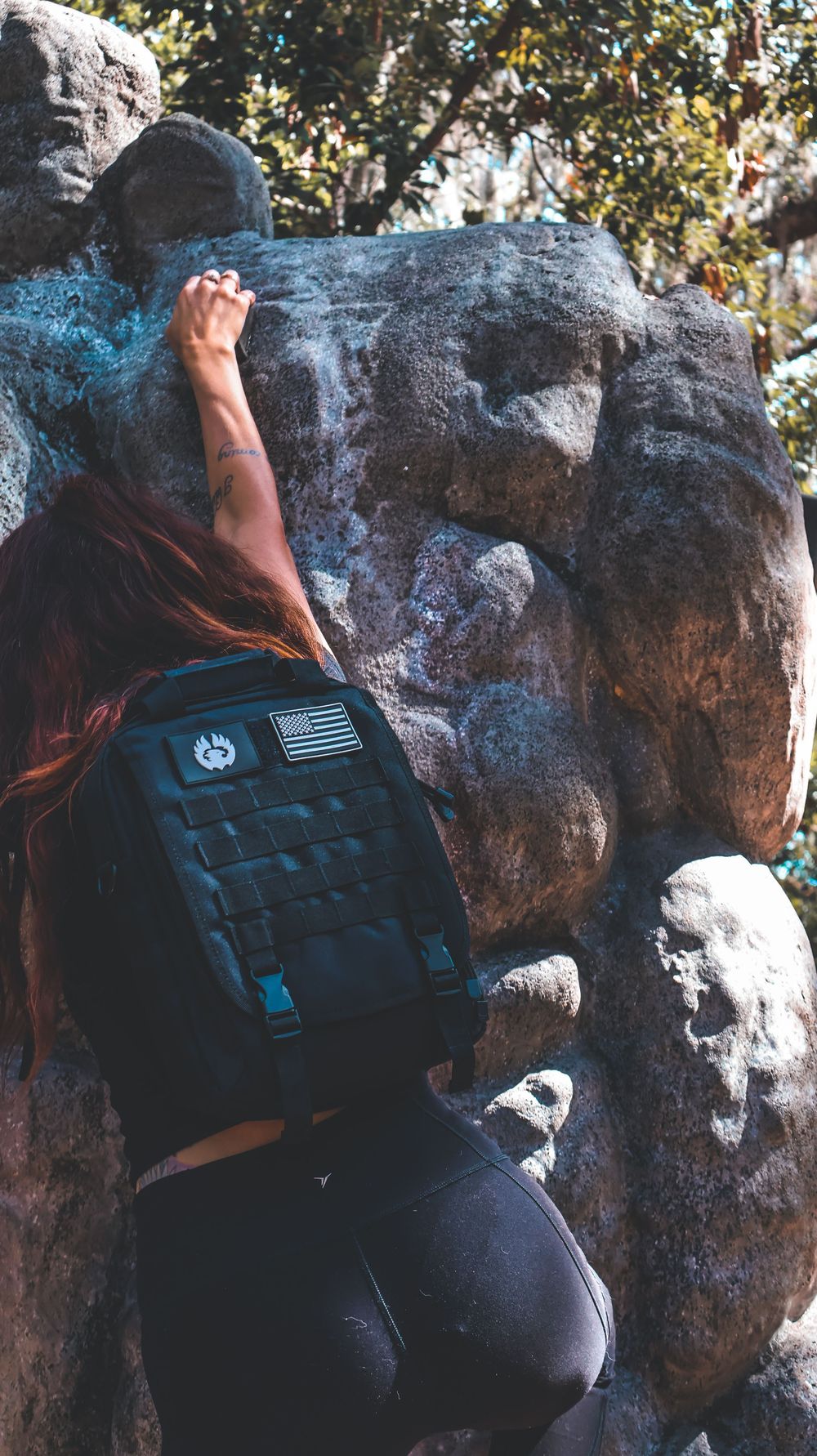 Woman with red hair and black tactical backpack rock climbing on large boulders outdoors.