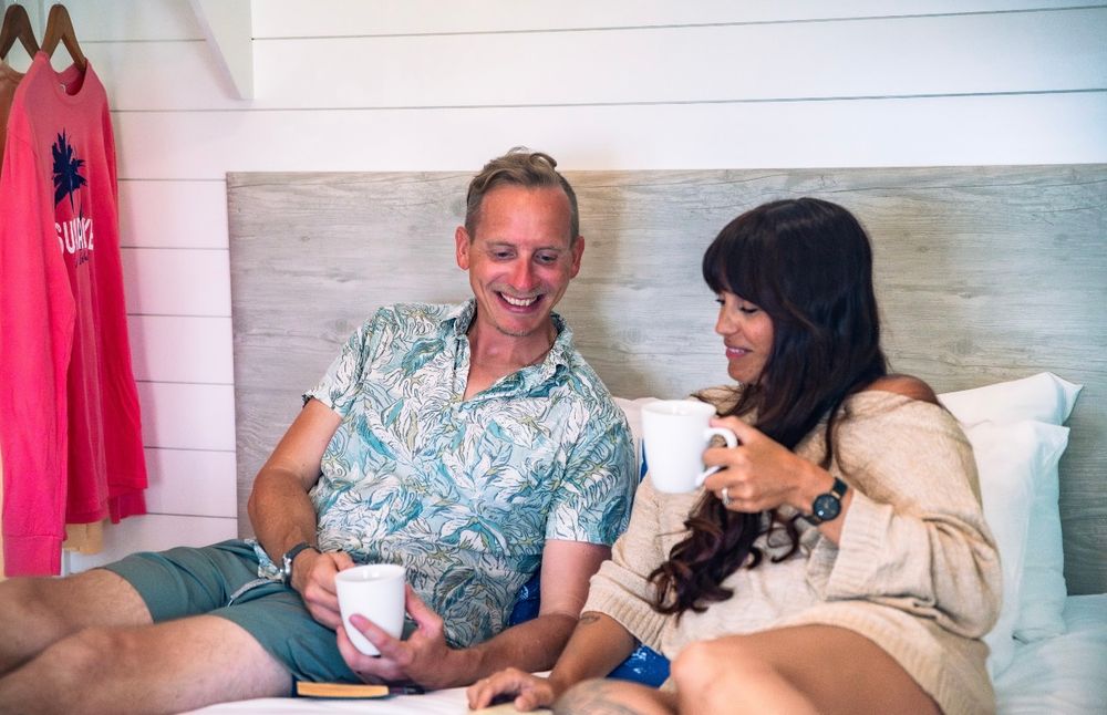 Smiling couple relaxing in bed with coffee cups in a cozy beach cottage bedroom.