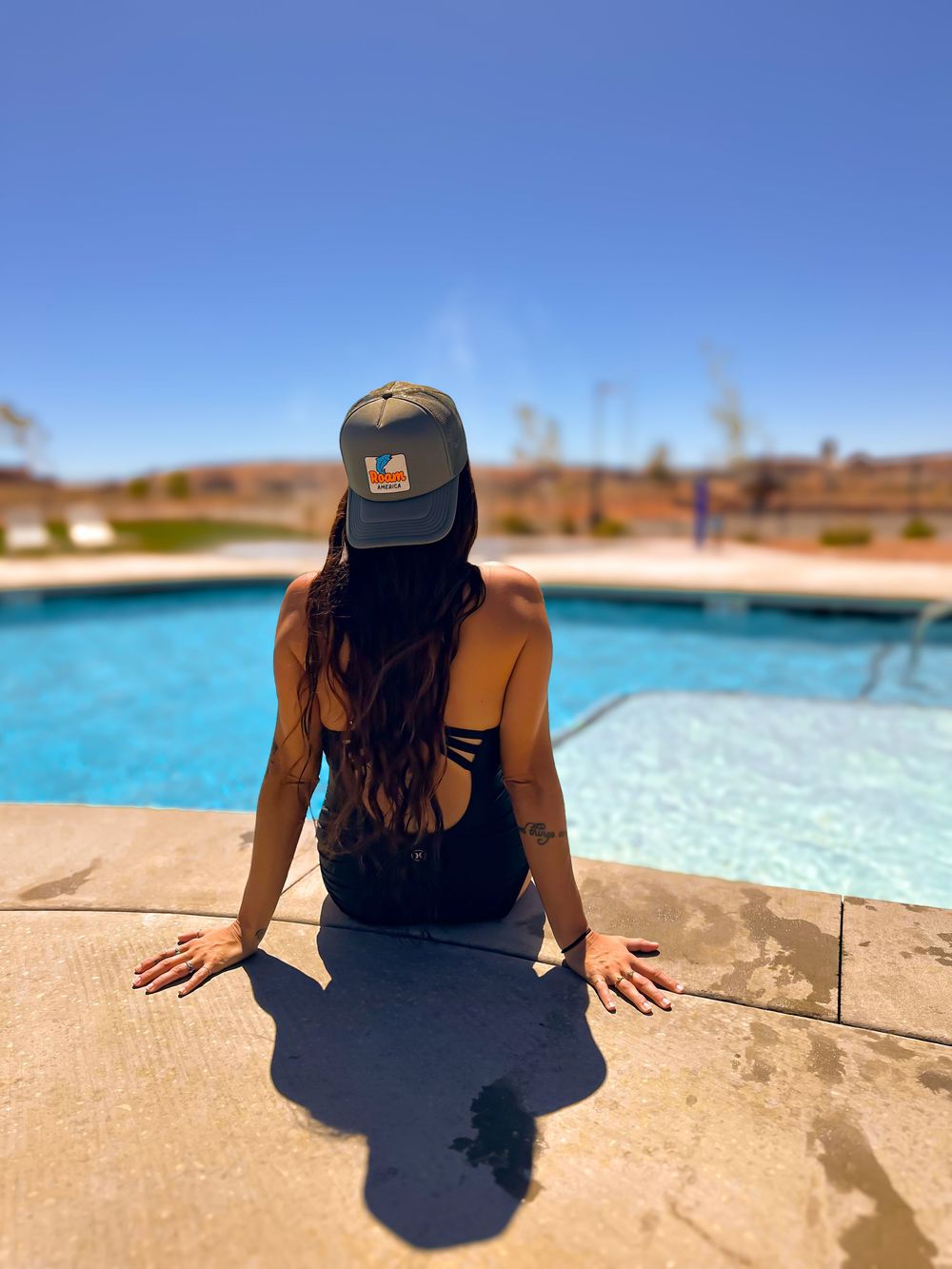 Woman in black swimsuit and gray cap sitting poolside in sunny desert resort.