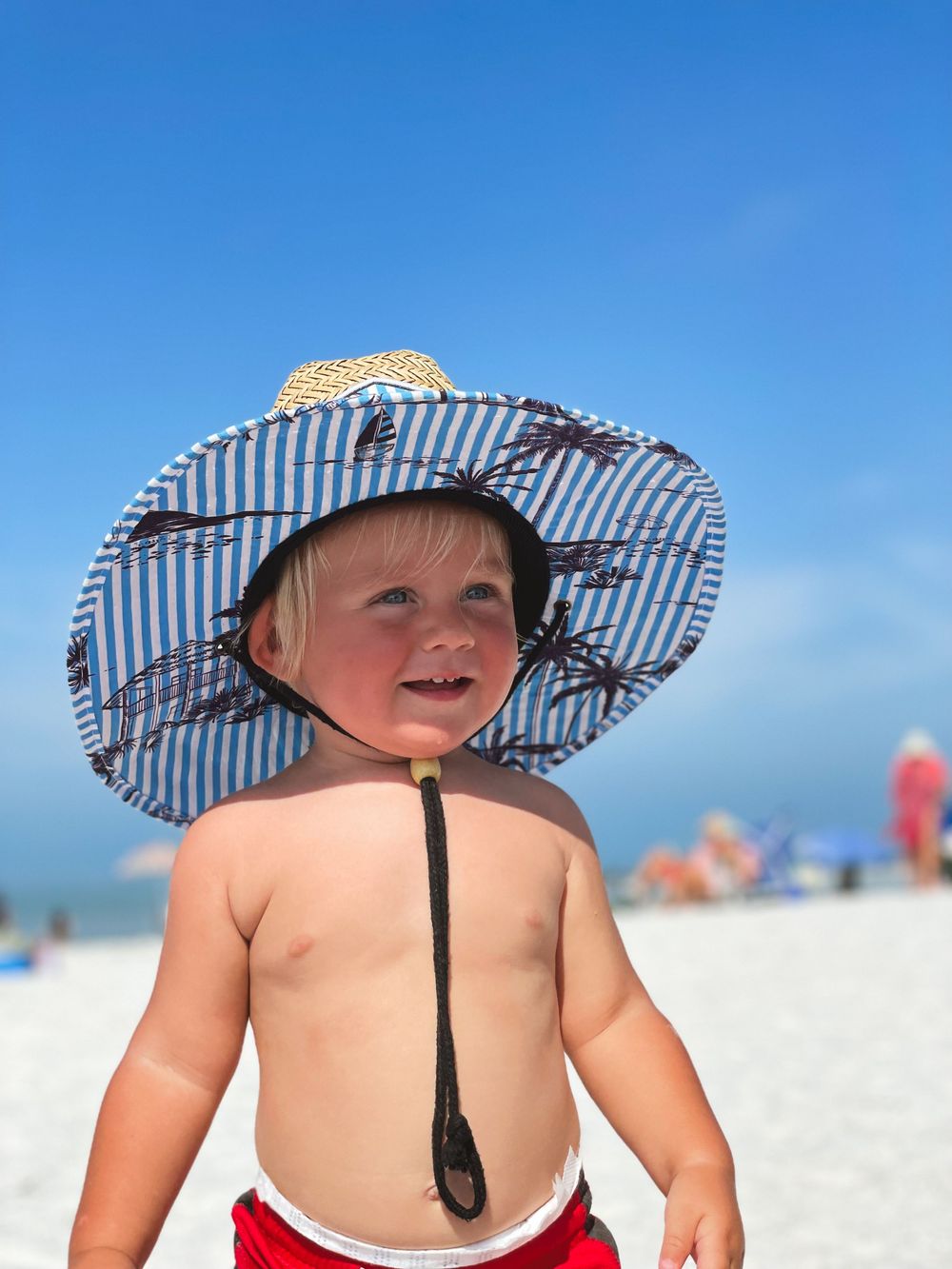 Adorable toddler wearing oversized striped sun hat on a sunny beach with blue sky.
