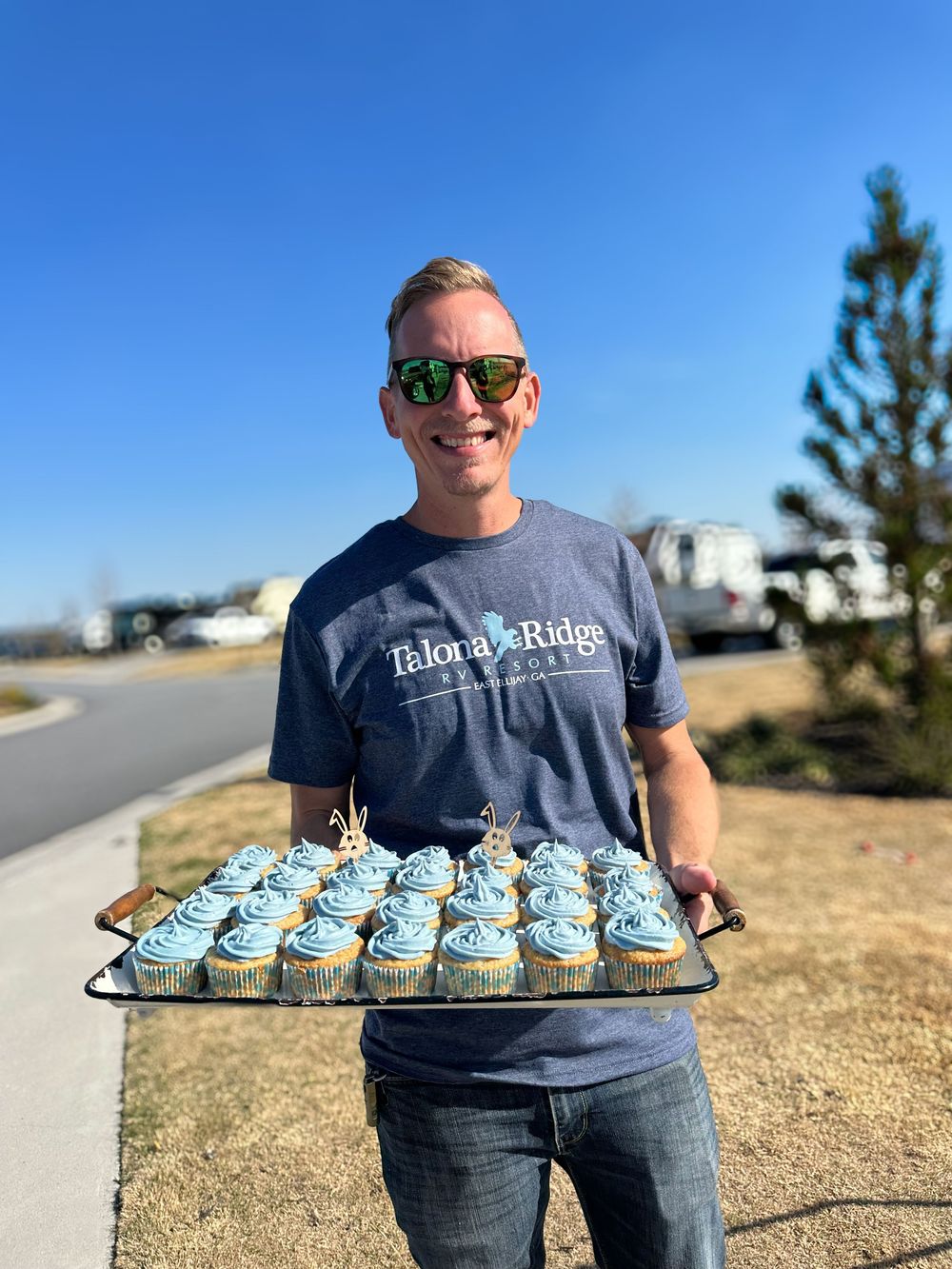 Smiling man holding tray of blue frosted cupcakes outdoors at Talouga Ridge RV resort.