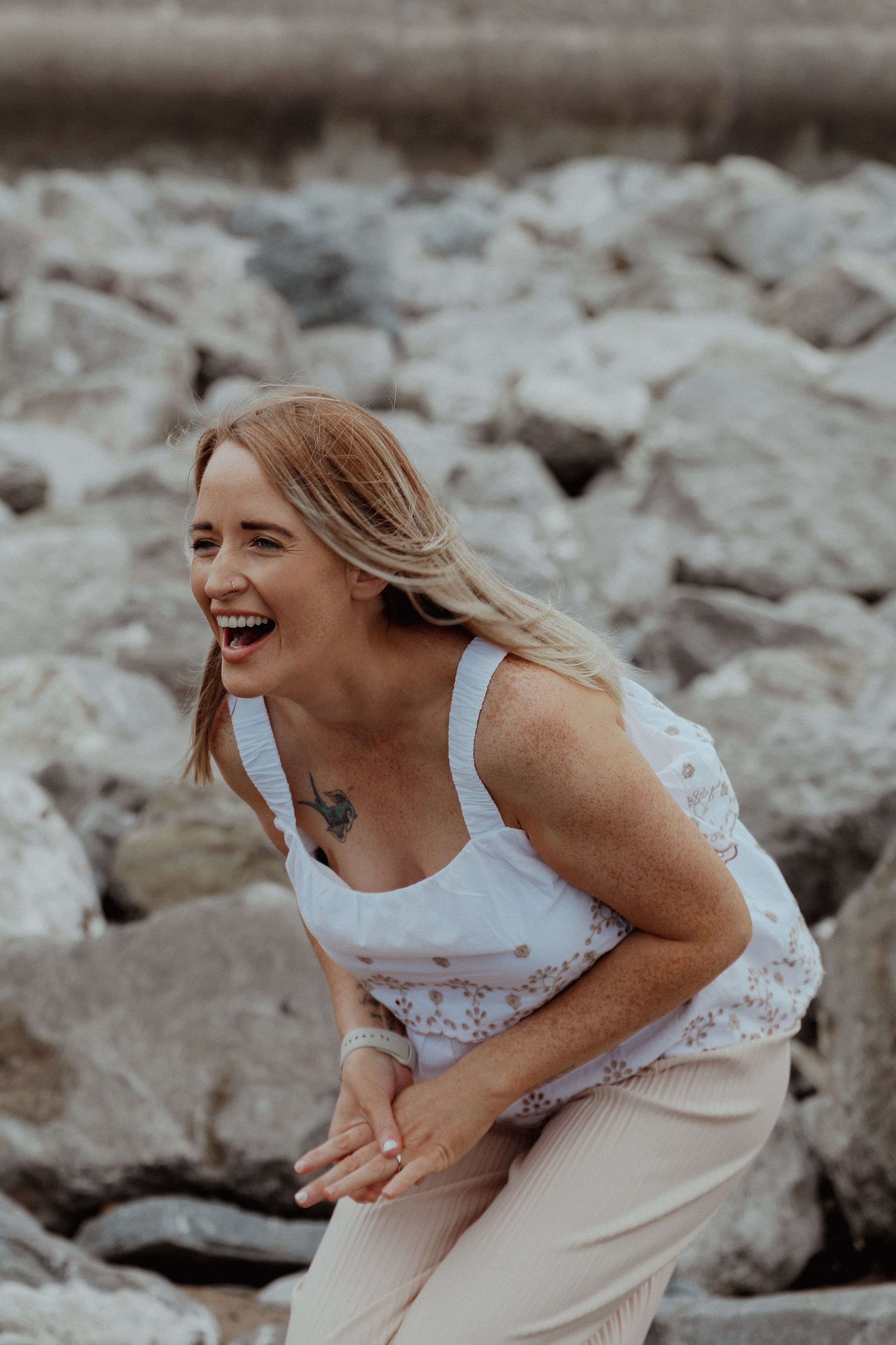 A woman in a white tank top and beige pants laughs while crouching on rocky terrain.