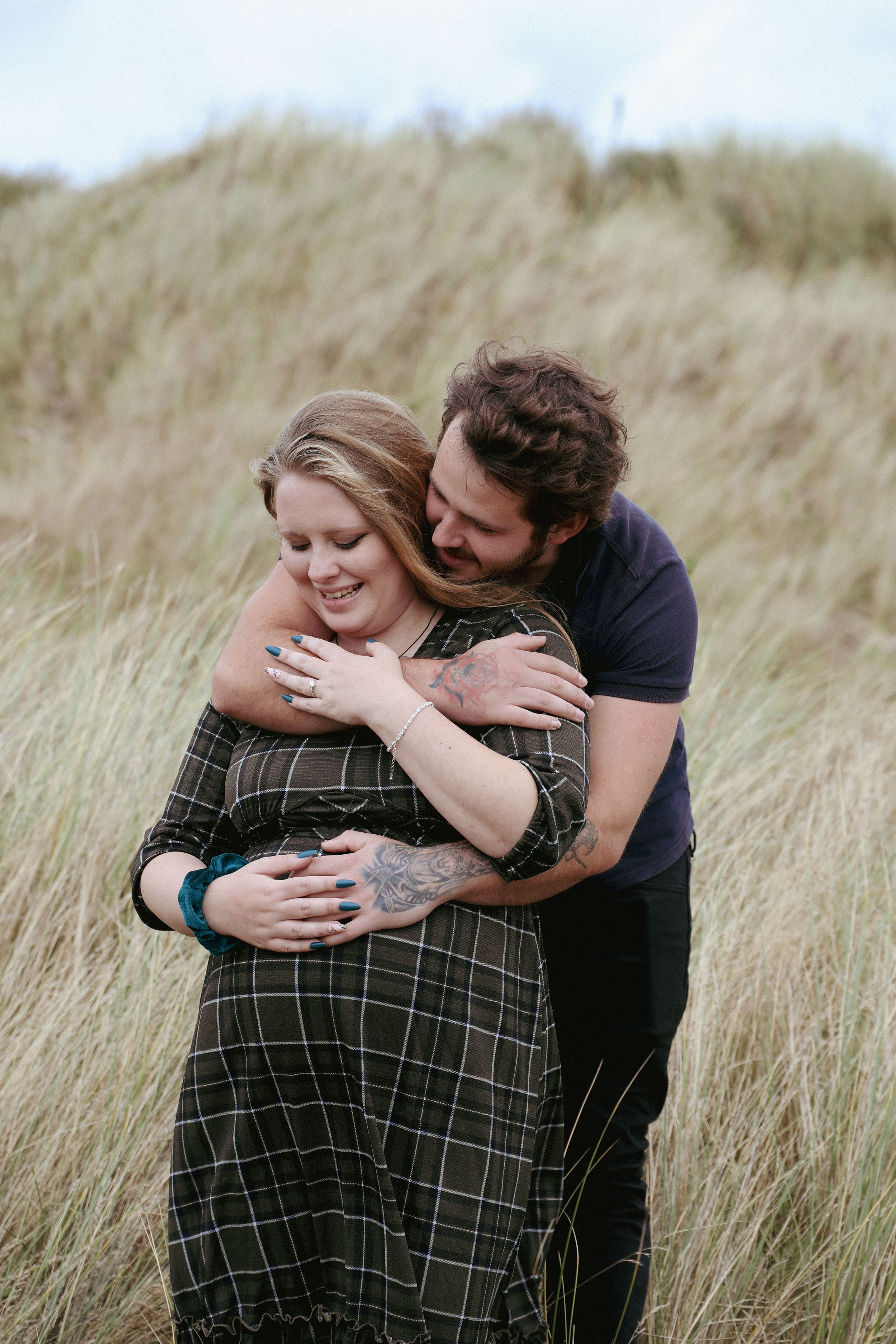 A couple shares an intimate embrace while sitting together in tall windswept grass during an outdoor photo session.