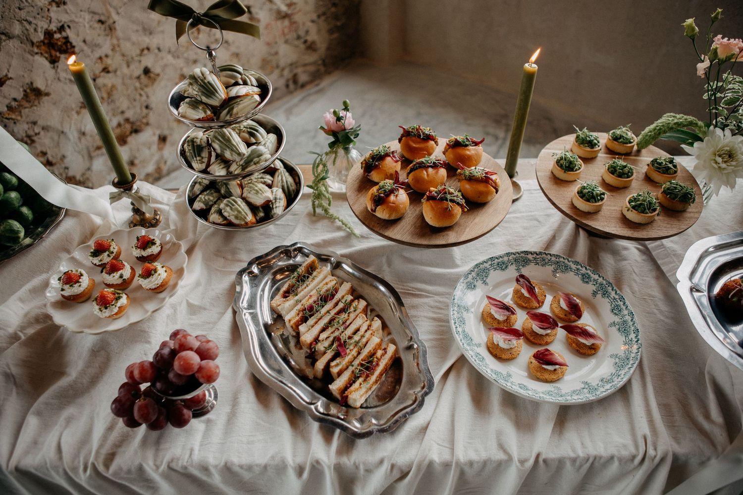 Wedding grazing table with sandwiches, stuffed mushrooms, fruit tarts, and tiered dessert stand on white linen.