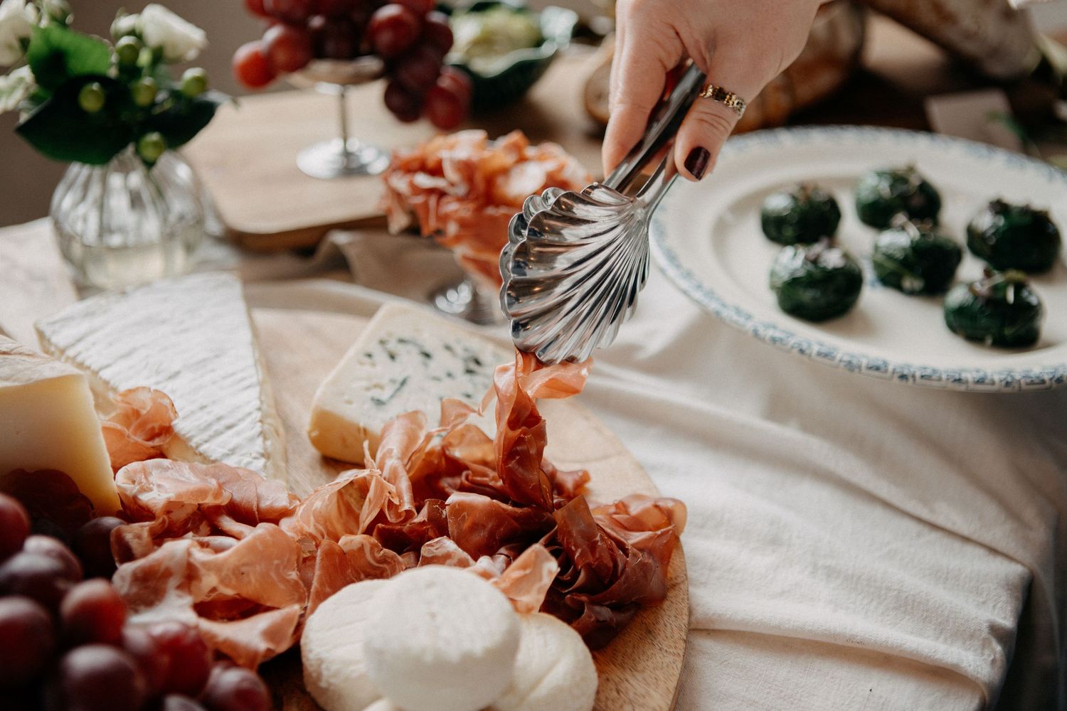 Hand using tongs to serve from a charcuterie board with prosciutto, cheese, and mozzarella at a wedding reception.