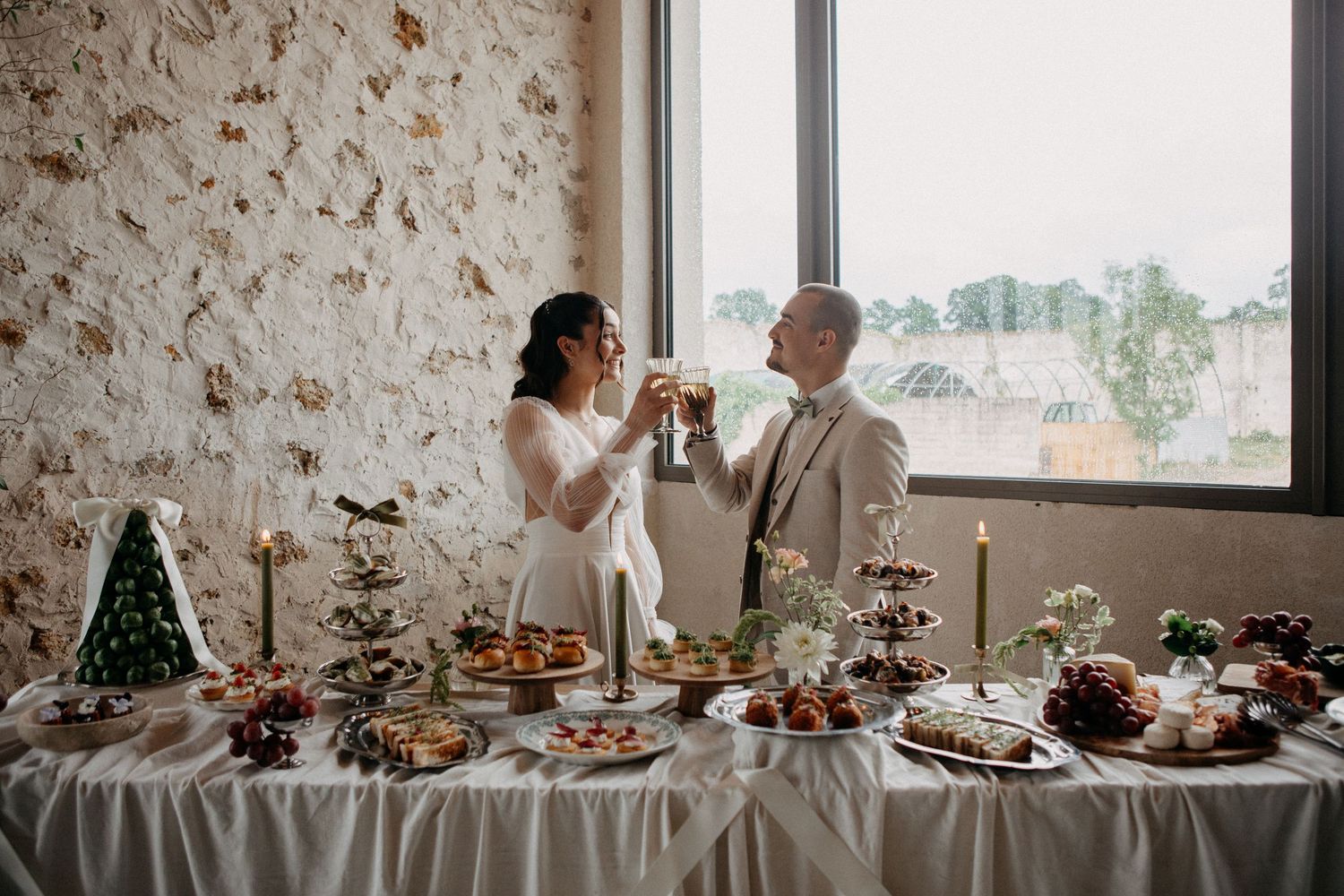 Bride and groom toasting champagne at an elegant wedding dessert table with candles and treats by a large window.