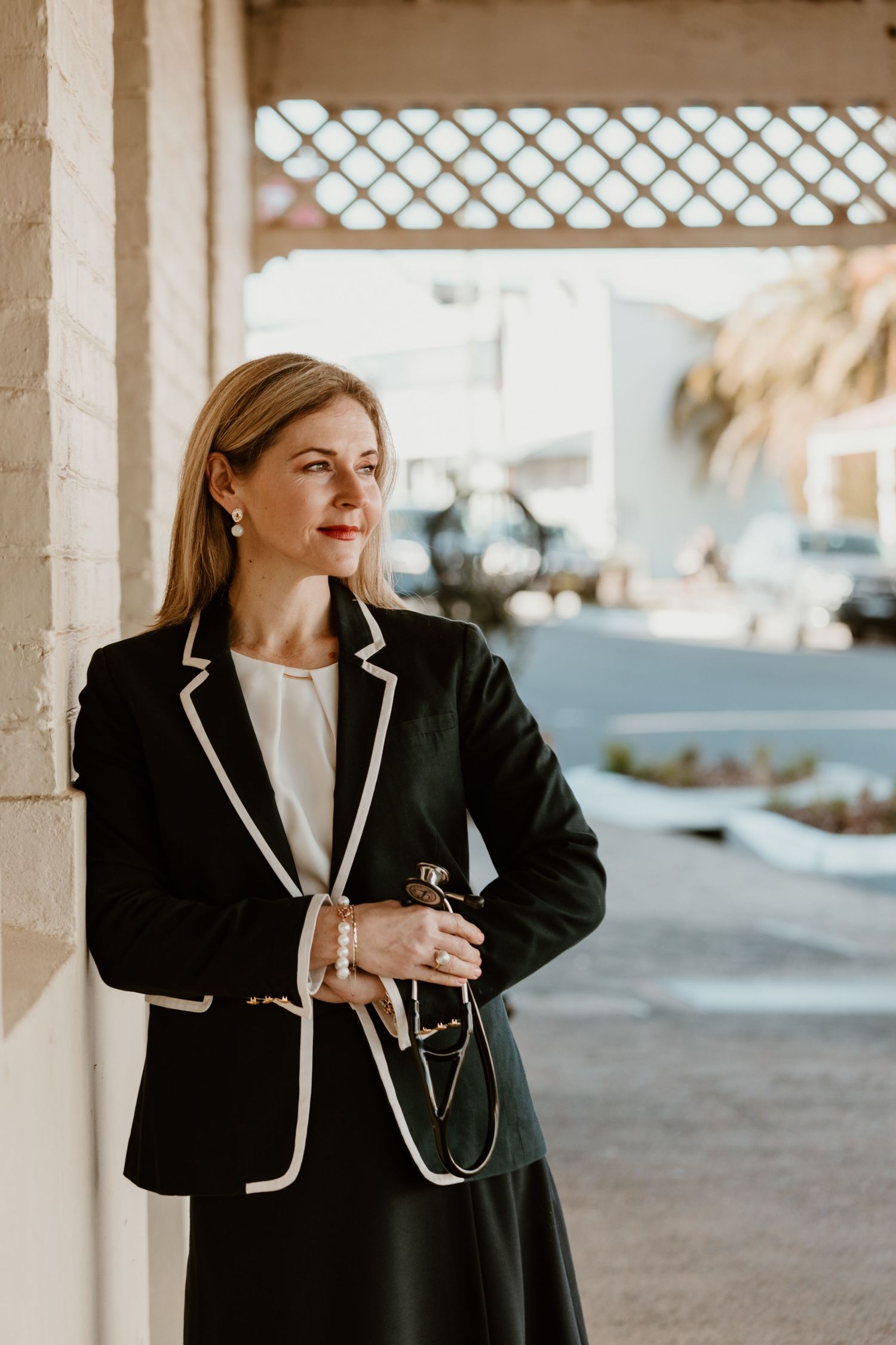 Professional wearing a classic black blazer with white trim stands confidently against a beige wall in an outdoor setting.